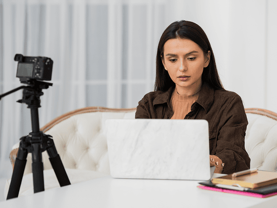 Woman researching mental health resources on a laptop, camera on tripod nearby.
